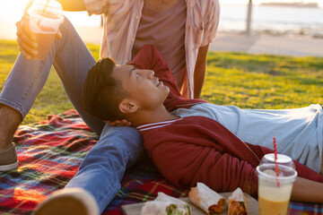 Happy biracial gay male couple having picnic on promenade by the sea at sundown