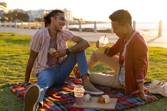 Happy biracial gay male couple having picnic on promenade by the sea at sundown