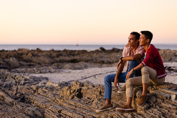 Happy biracial gay male couple sitting on rocks and embracing on beach at sundown, with copy space