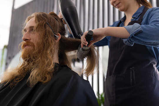 Caucasian female hairdresser styling long hair of smiling male customer with hairdryer at hair salon