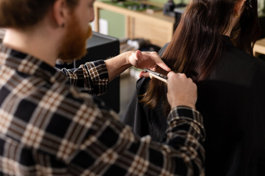 Focused bearded caucasian male hairdresser cutting long hair of female customer at hair salon