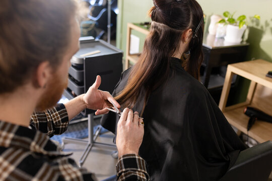 Focused caucasian male hairdresser cutting ends of female customer's long hair at salon