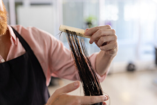 Midsection of caucasian male hairdresser combing long hair of customer at hair salon