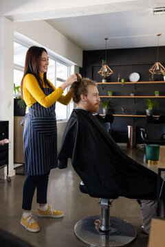 Happy caucasian female hairdresser undoing hair bun of long haired male client at hair salon