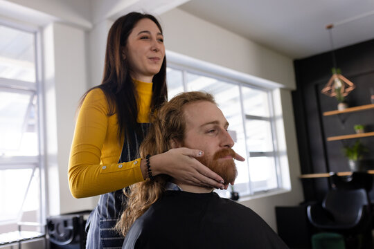 Happy caucasian female hairdresser holding beard of long haired male client at hair salon