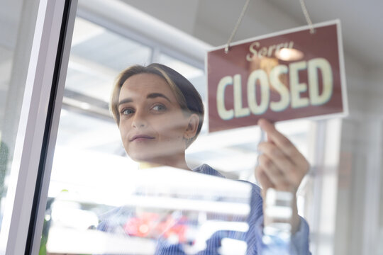 Happy caucasian female hairdresser turning sign to closed on glass door of hair salon
