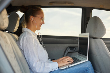 Side view portrait of confident woman working on laptop while sitting in the car listening music while riding to work smiling typing on keyboard freelance job.