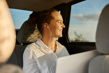 Beautiful enjoyable woman wearing white shirt working on laptop while sitting in the car and...