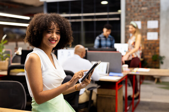 Portrait of happy biracial businesswoman with tablet over diverse colleagues in office, unaltered