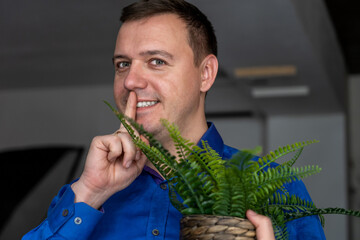 Mysterious attractive guy with fern house plant showing shh sign in indoors. Millennial man holding...