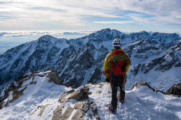 Adventurous man hiker on top of a steep rocky cliff overlooking winter alpine like moutain landscape of High Tatras, Slovakia. Alpine mountain landscape covered with glaciers, snow and ice.