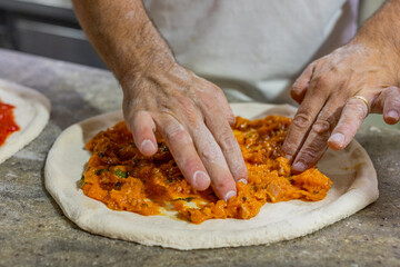 Pizzaiolo napoletano mentre stende con le mani una crema di zucca su un impasto di pizza napoletana crudo su un banco da lavoro in marmo di una pizzeria 