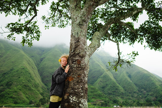 A young, beautiful girl hugs a tree in nature, against the backdrop of green mountains and enjoys life.