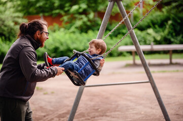 Obraz premium Little toddler boy happily rides on swings and playing with tall long haired man in glasses