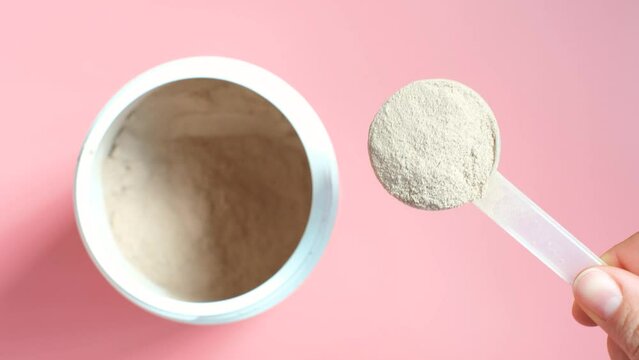 Female hand holding a scoop with chocolate protein next to jar on a pink background top view.