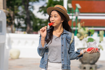 Portrait of Asian female traveler eating a slice of water melon while walking on sidewalk of buddhist temple on street in Bangkok, Thailand, Southeast Asia - Woman enjoying street food lifestyle - 605177724