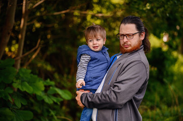 Young long hair bearded man in glasses holding little toddler boy on arms in green parks