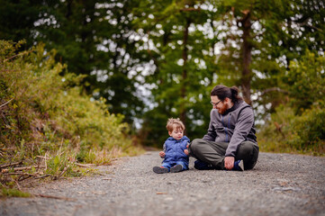 Fototapeta na wymiar Young long hair bearded man in glasses sitting with little toddler boy on road playing with stones