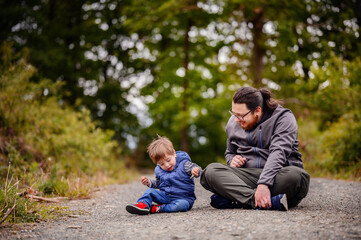 Young long hair bearded man in glasses sitting with little toddler boy on road playing with stones