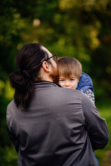 Young long hair bearded man in glasses holding little toddler boy on arms in green parks