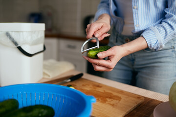 Housewife peeling a cucumber skin with a peeler tool in a domestic kitchen. Female person preparing a healthy green salad at home and recycling organic waste in a compost bin. Sustainability concept