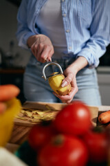 Housewife cooking vegetables at home. Female person peeling off a a potato with a peeler tool in a domestic kitchen in the morning
