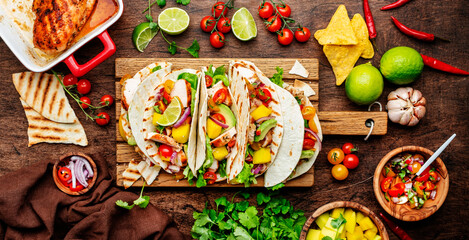 Taco party. Corn tortillas with grilled chicken fillet, salsa sauce, mango, cilantro and red onion on rustic wooden cutting board. Wood table background, top view
