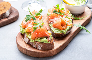 Avocado salmon sandwich on rye bread with guacamole sauce, young arugula and sesame seeds on wooden cutting board, gray table background, top view