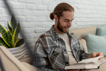 Happy young man reading book while sitting on sofa in his living room.