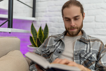 Happy young man reading book while sitting on sofa in his living room.