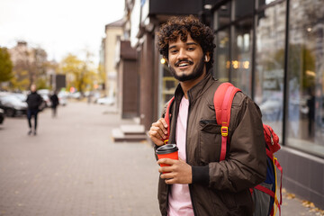 Smiling indian tourist with backpack and coffee to go looking at camera on city street