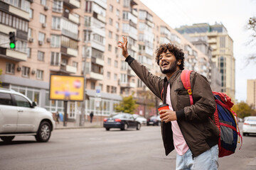 Positive indian backpacker holding coffee and catching taxi on road in city 