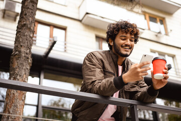 Young smiling indian man in earphone using smartphone and holding coffee on urban street 