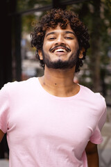 Portrait of young and curly indian man in pink t-shirt looking at camera and smiling while standing on blurred urban street
