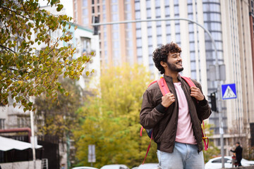 Smiling young indian man holding backpack while traveling and standing on blurred urban street at background