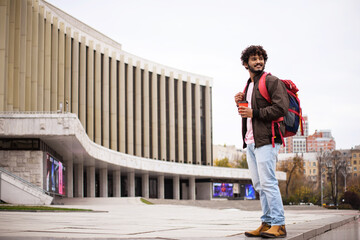 Positive young indian man in casual clothes holding backpack and coffee to go while standing on blurred urban street 