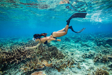 A young woman in bikini free dives above a coral reef in clear tropical water	