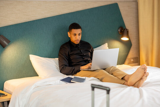 Young dark-haired man lying on bed in a hote room and surfing internet