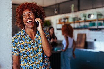 Young African-American man laughing during phone call while two women talk in the background. - obrazy, fototapety, plakaty