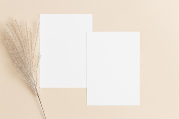 Two white invitation cards mockup with a reed pampas decoration on the beige table. 5x7 ratio,...