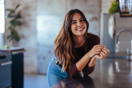 Portrait of young adult woman smiling while leaning on counter in kitchen.