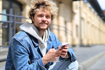 Curly-haired blond man is sitting with phone outside.