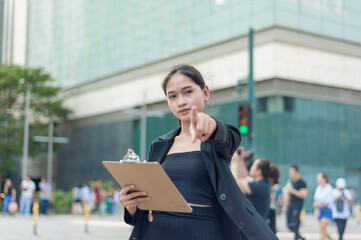 A lovely lady standing outside wearing a black suit, and black skirt seriously looking and pointing at the camera while holding a clipboard. A building, traffic light and people in the background.