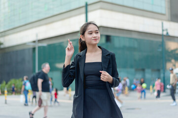 A lovely lady standing outside, in the city wearing a black suit and black skirt holding up a crossed finger, smiling while gazing far. A building, traffic light and people in the background.