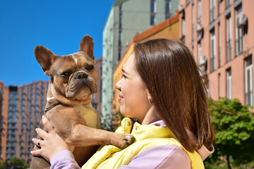 Portrait affectionate woman spending leisure with French Bulldog at public park. Caring young pet owner showing tenderness small dog outdoors. Colorful buildings of residential district on background
