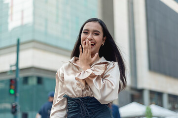 A pretty asian woman standing outside wearing a light tan silk blouse covering her laugh while looking at the camera. A building in the background