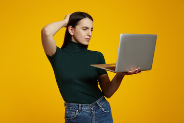 Portrait of a doubtful young girl holding laptop computer, wearing green t-shirt while standing isolated over yellow background.