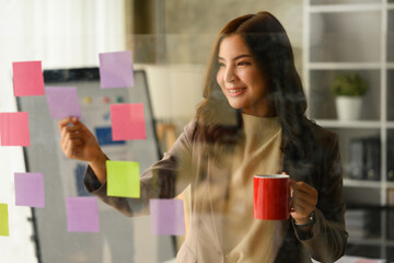 Successful startup founder drinking coffee and reading sticky notes on glass wall, analyzing sales...