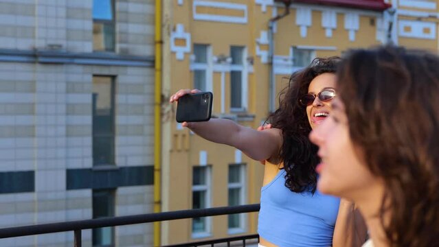 Two friends using mobile phone while taking photo, standing on the rooftop while their friends dancing. Lifestyle, fun concept. Real time