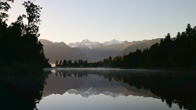 Serene, misty sunrise over Lake Matheson, New Zealand; snowy mountain peaks reflecting in calm water